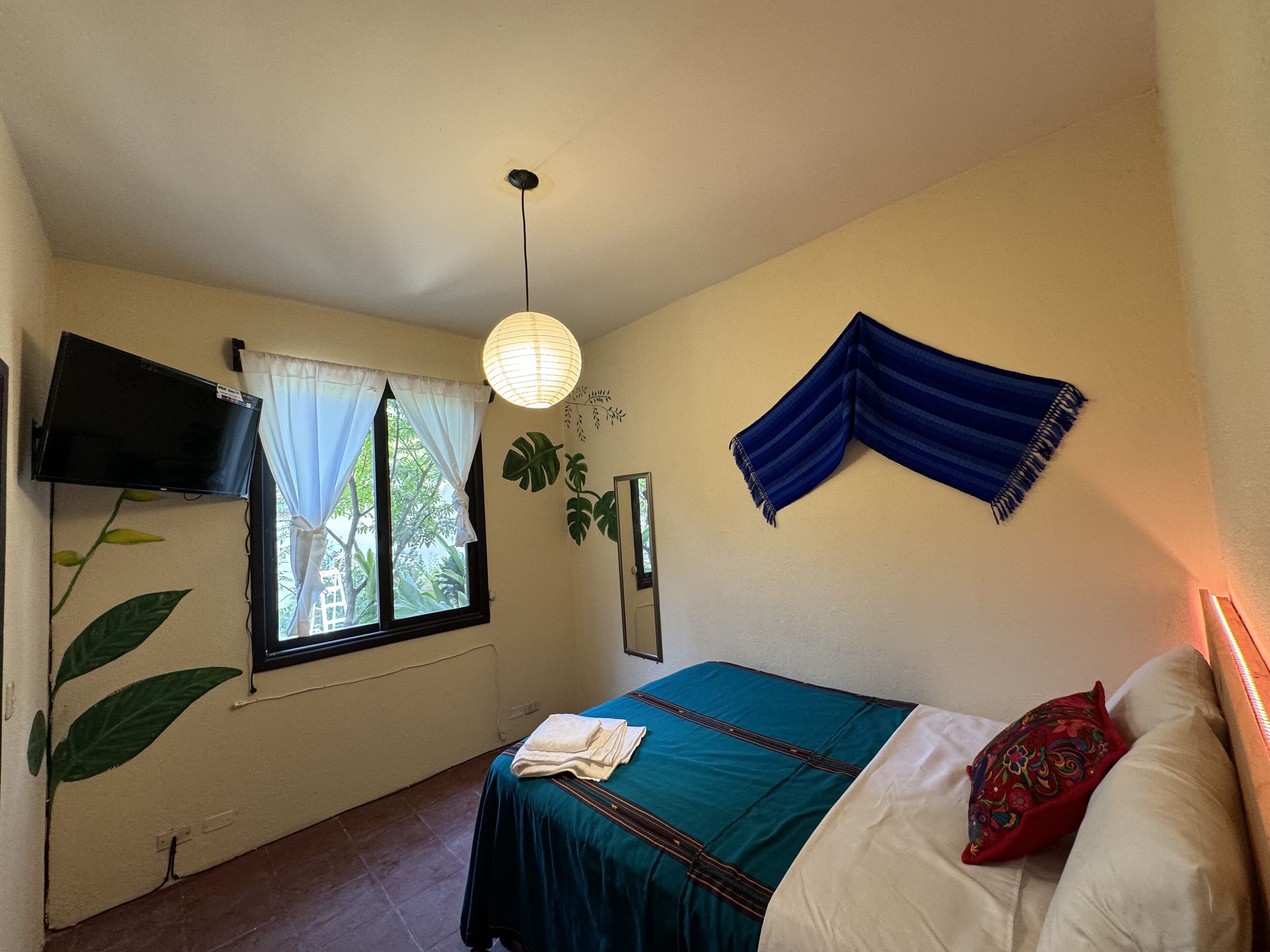 Un dormitorio con cama, televisión montada en la pared, ventana con cortinas y una planta colgante.