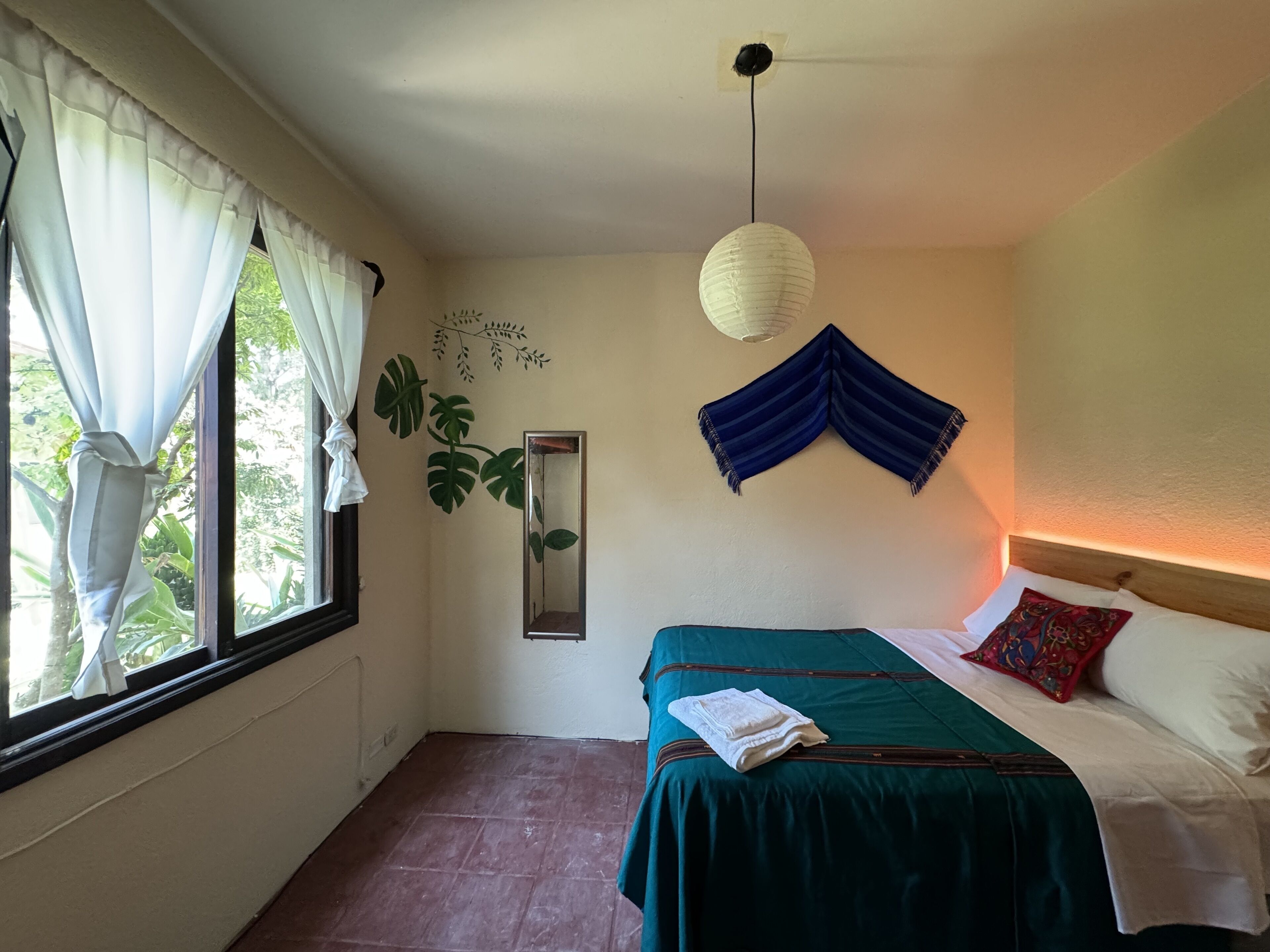 Un dormitorio con cama, ventana con cortinas, lámpara colgante y planta en la pared.