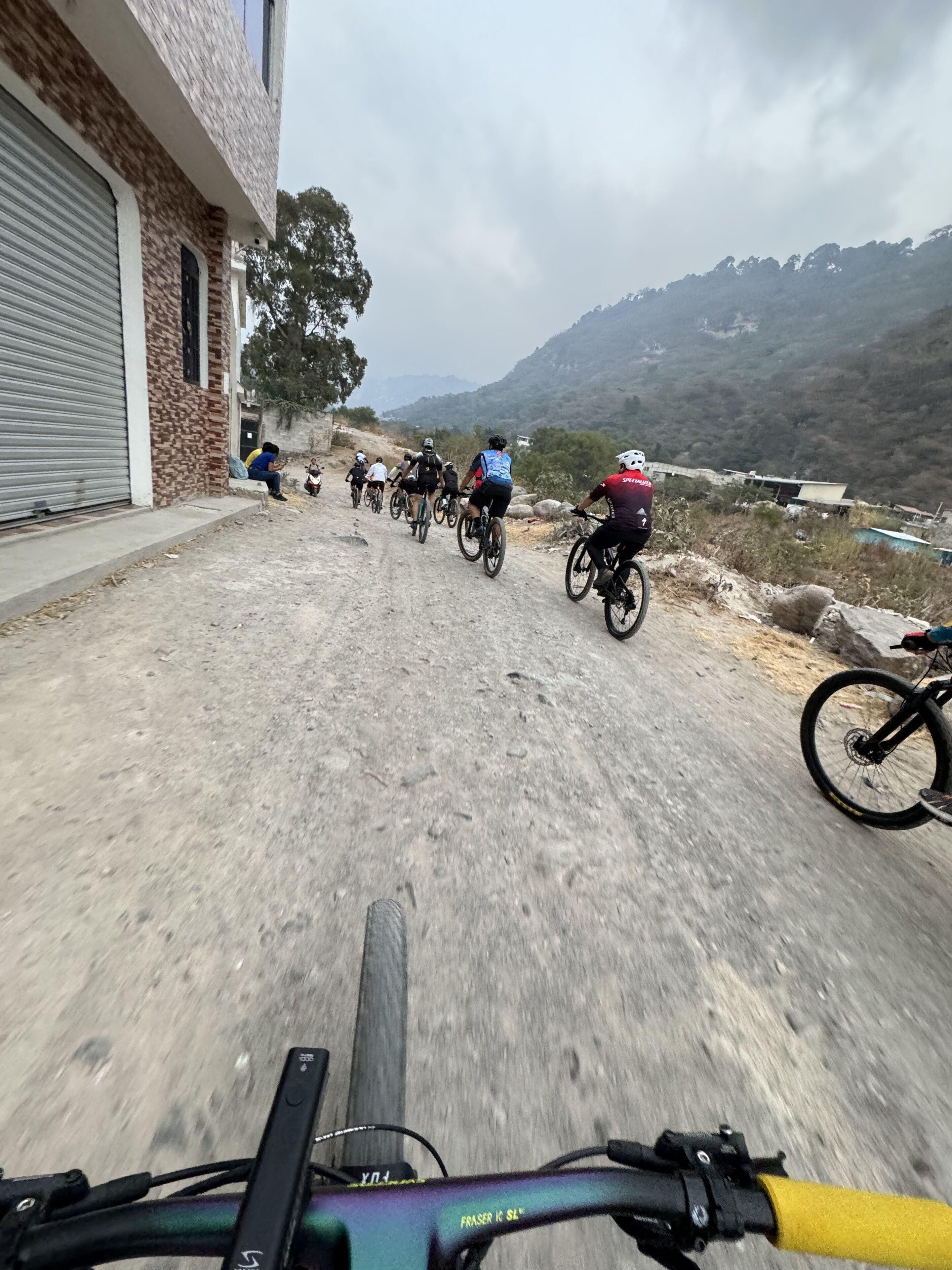 Un gruppo di ciclisti su un sentiero sterrato, con un edificio a sinistra e montagne sullo sfondo.