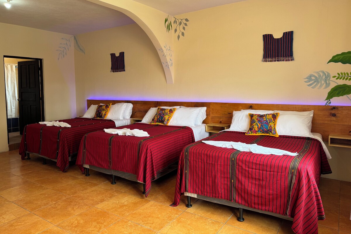 Ein Hotelzimmer mit drei Betten, jeweils mit rot gestreifter Bettwäsche und weißen Kissen. Es gibt ein hölzernes Kopfstueck, einen kleinen Nachttisch aus Holz und eine Wand mit Blatt- und Pflanzendekorationen.