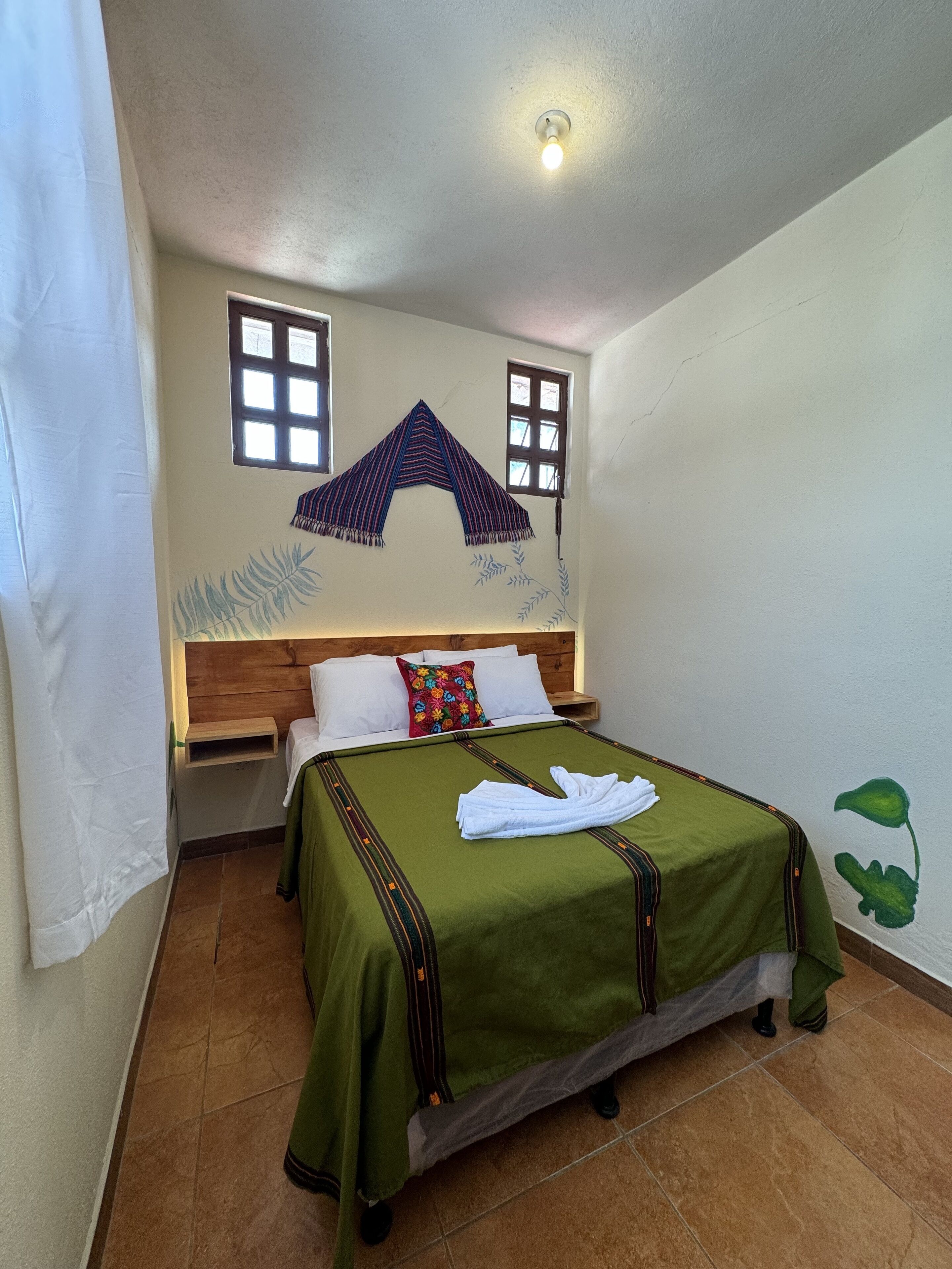 Een eenpersoonsbed met een groene dekbedovertrek, witte kussens en een rood kussen met bloemenprint. Er staat een klein houten nachtkastje naast het bed.