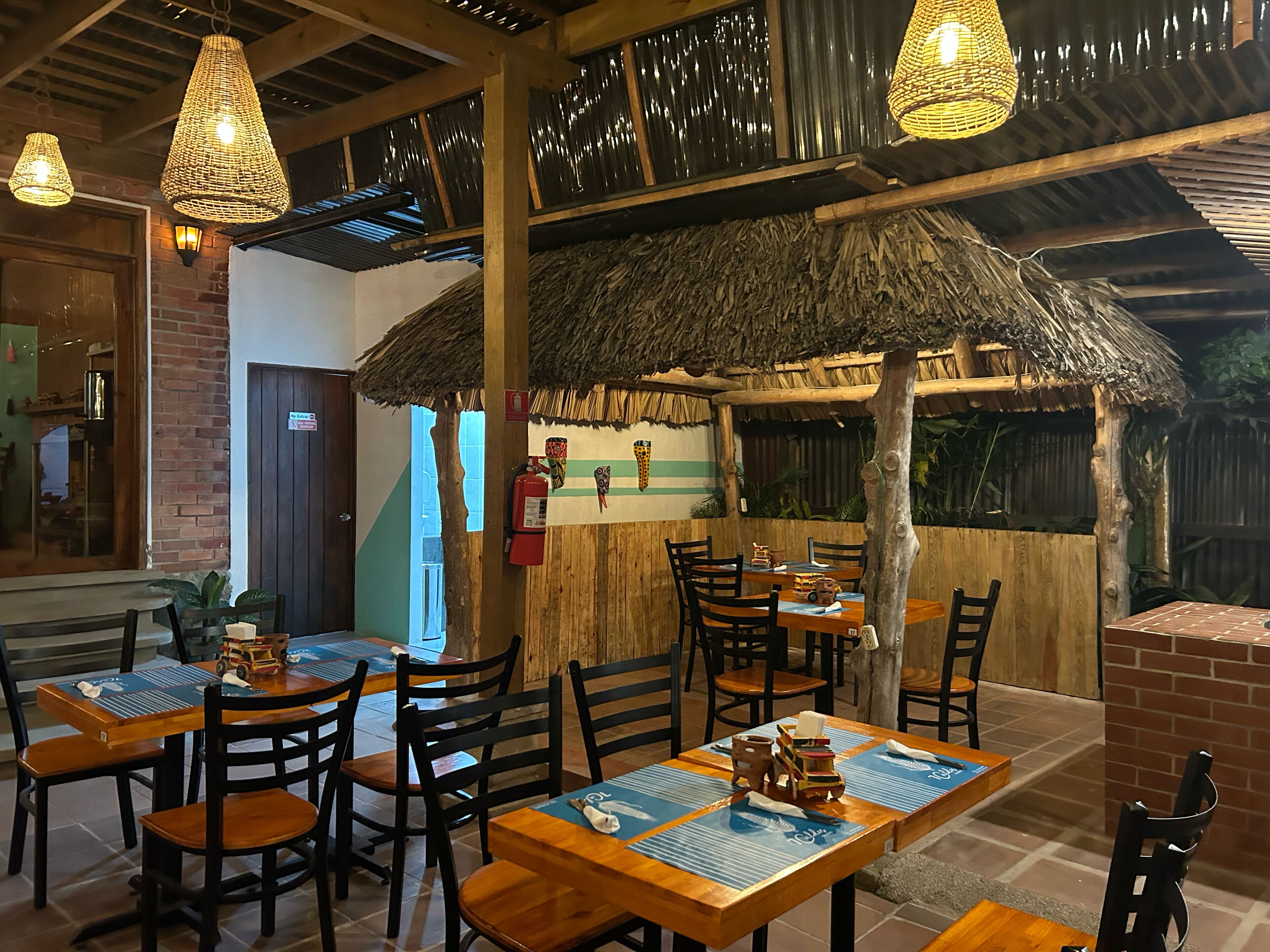 A restaurant with wooden tables and chairs, thatched-roof outdoor seating, and hanging wicker lamps.