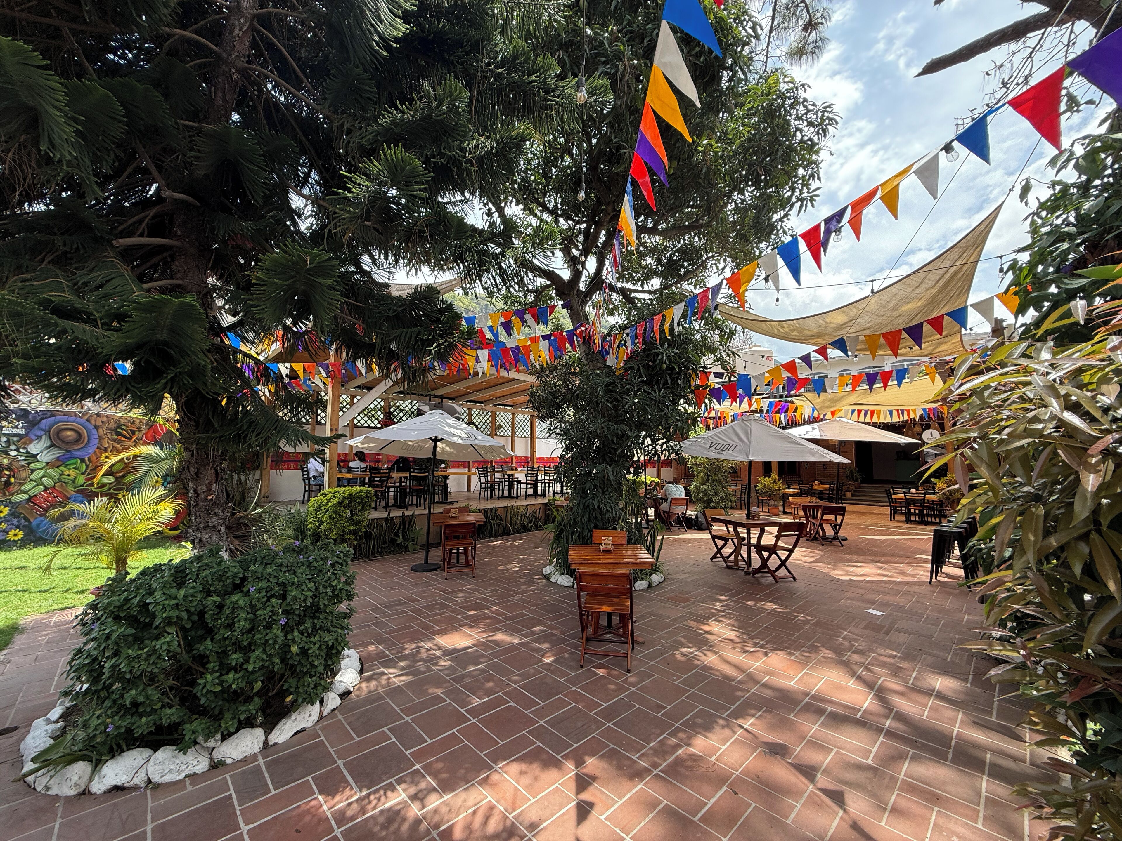 Un'area patio all'aperto con pavimento in mattoni, tavoli e sedie in legno e vivaci bandiere triangolari.