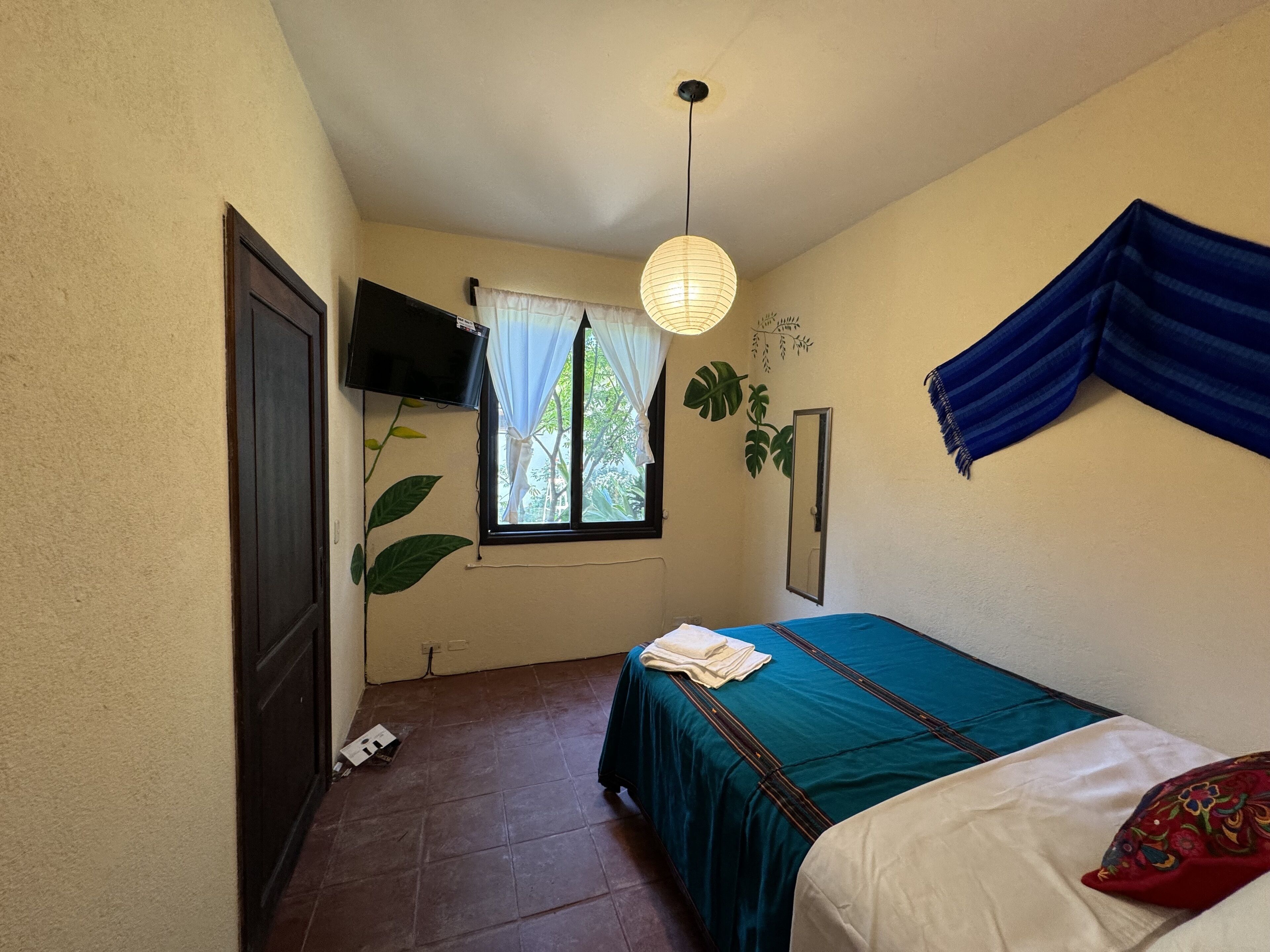Un dormitorio con una cama, un televisor montado en la pared, una ventana con cortinas y una planta.