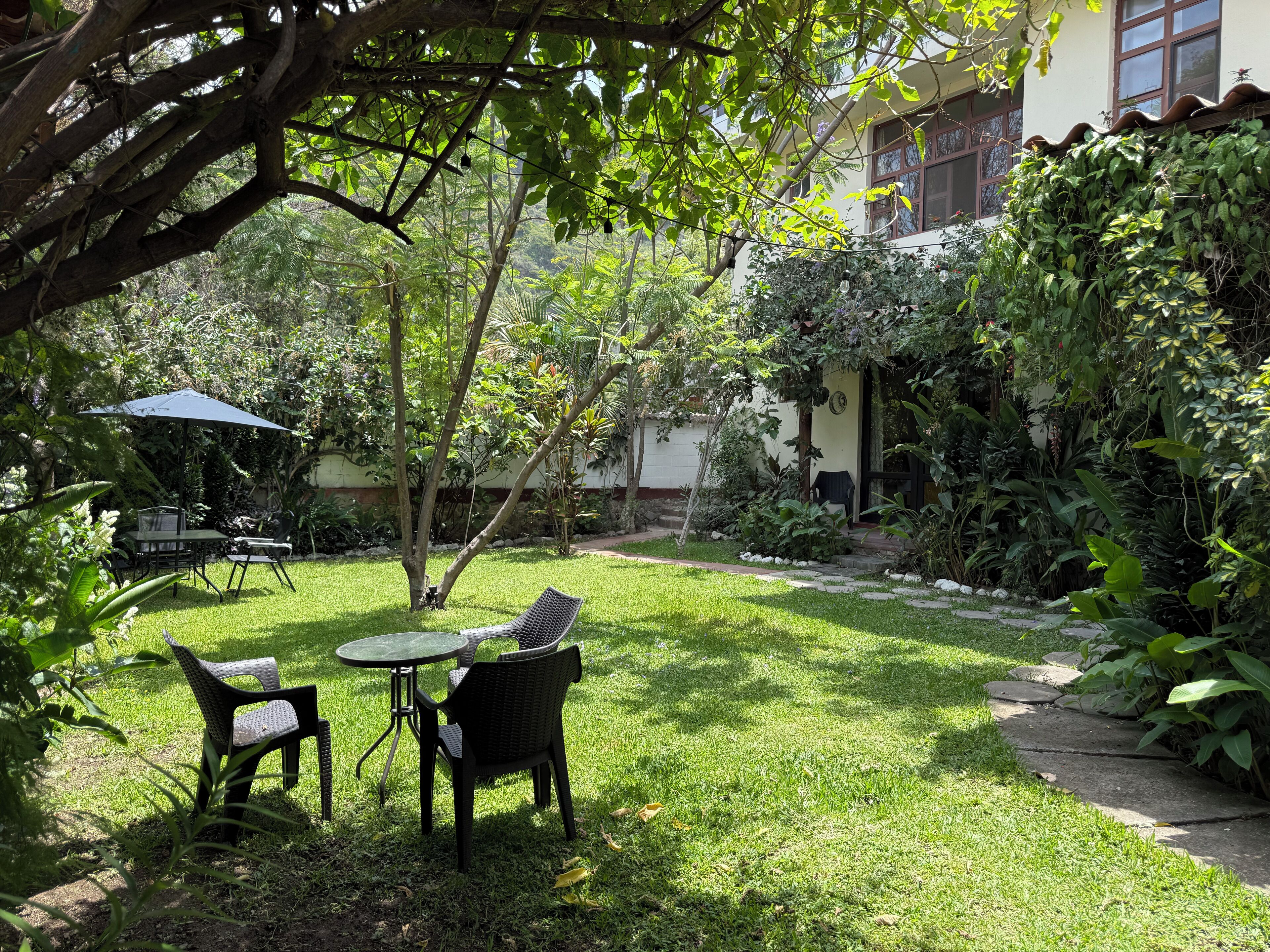 Un jardin avec une table et des chaises, entouré d’une végétation luxuriante et avec un bâtiment en arrière-plan.