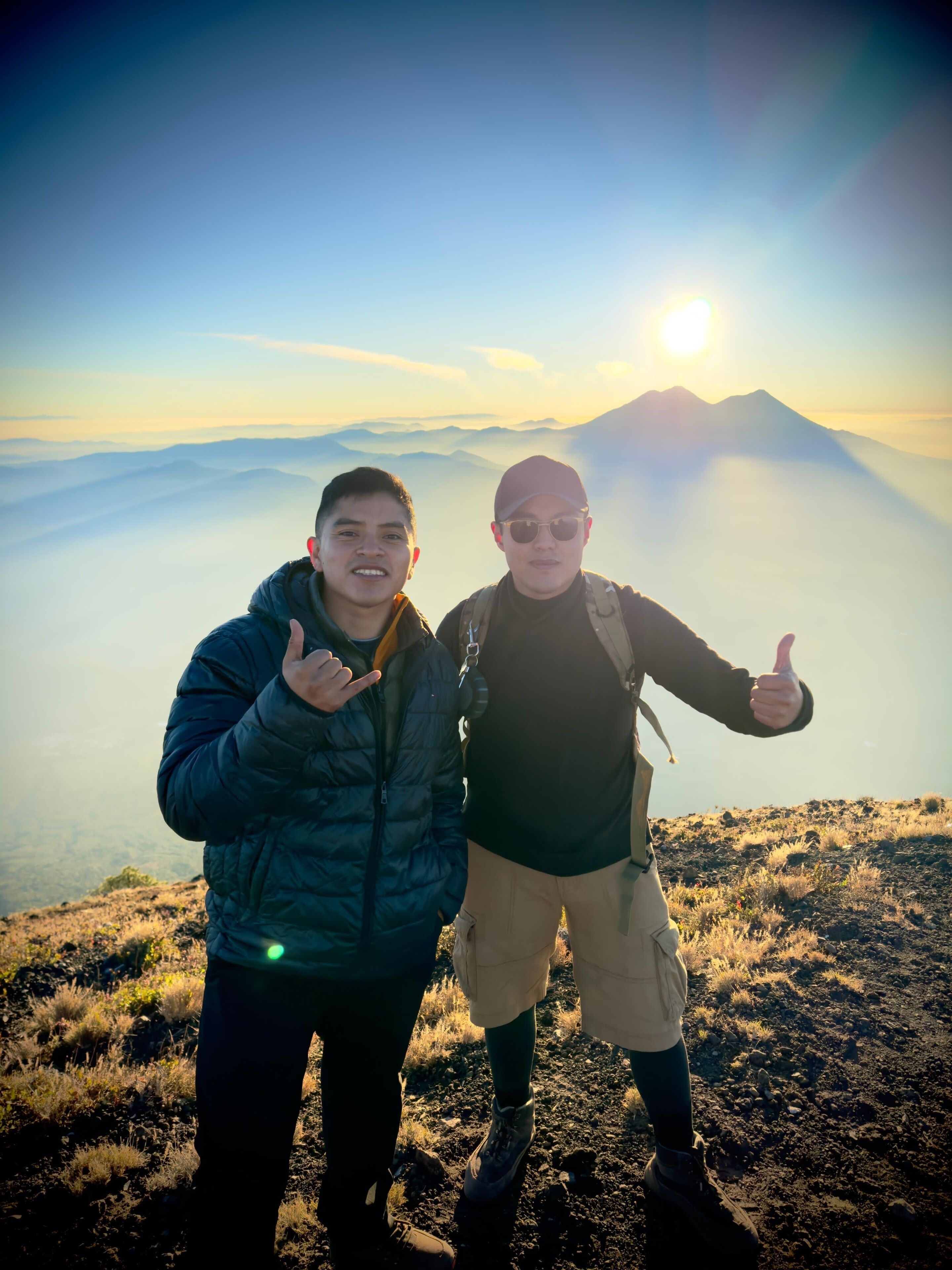 Dos personas de pie en una montaña con cielo despejado y sol al fondo.