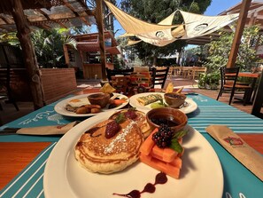 Une table à manger où sont disposés divers aliments pour le petit-déjeuner, notamment des crêpes, un saladier de fruits et une assiette avec des œufs et des toasts.