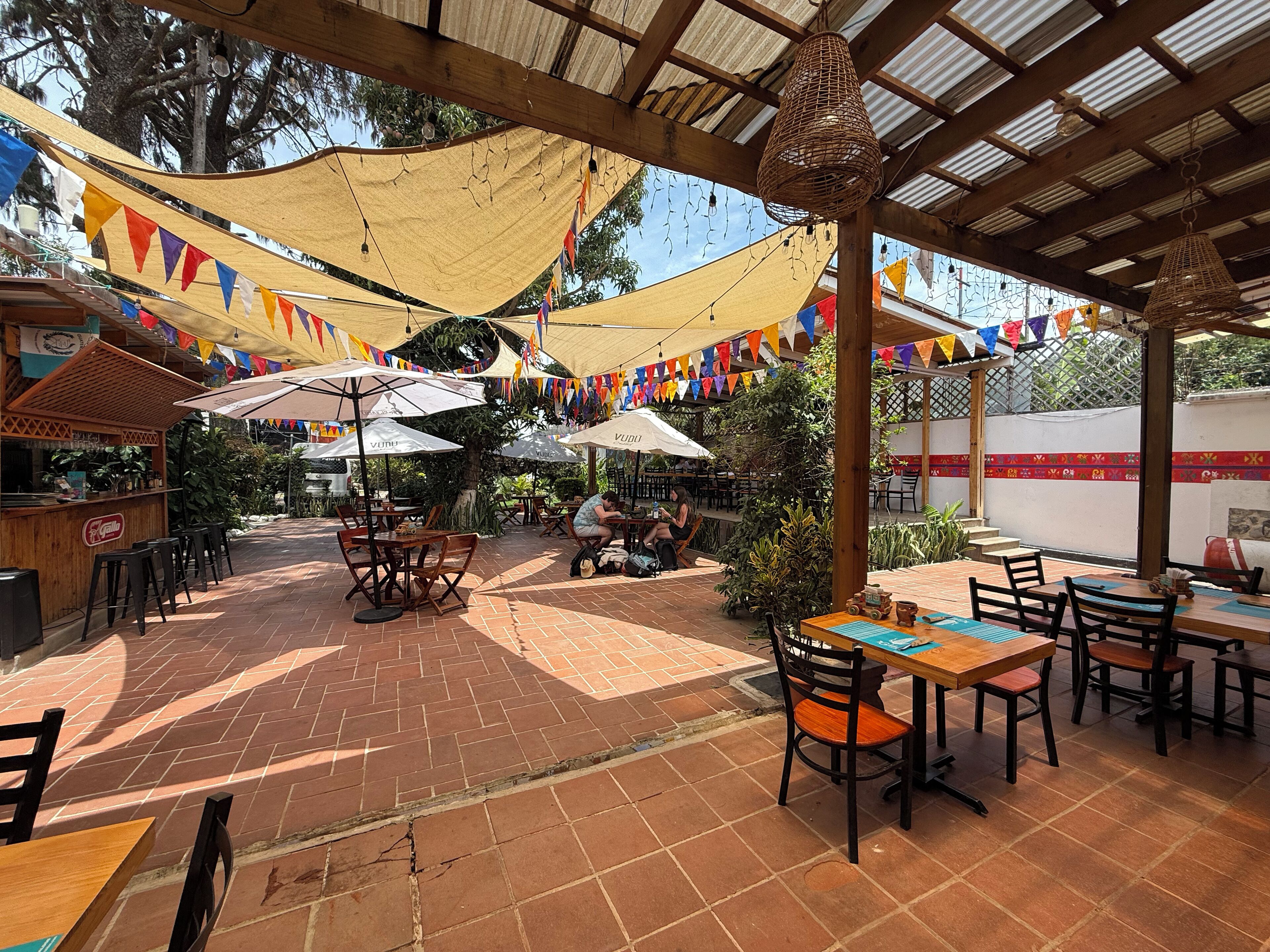 An outdoor patio area with tables and chairs, covered by a wooden pergola and shaded by a canopy.