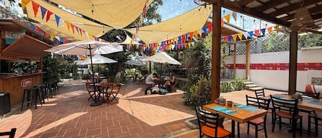 An outdoor patio area with tables and chairs, covered by a wooden pergola and shaded by a canopy.