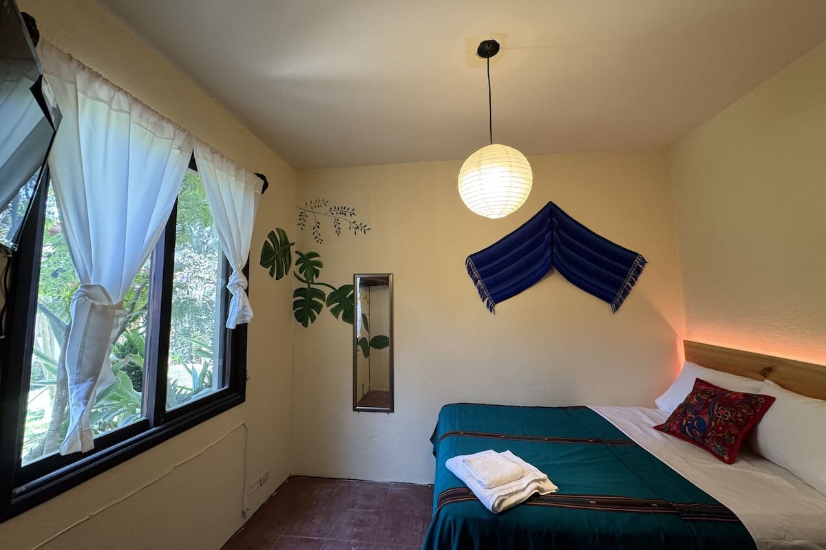 Une chambre à coucher avec un lit, une fenêtre avec des rideaux, un miroir fixé au mur et un luminaire suspendu.