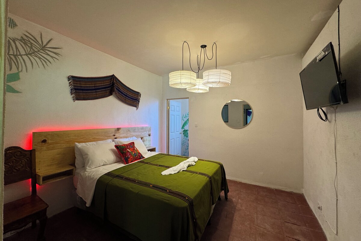 Een slaapkamer met een bed, nachtkastje, lamp en een aan de muur bevestigde televisie.