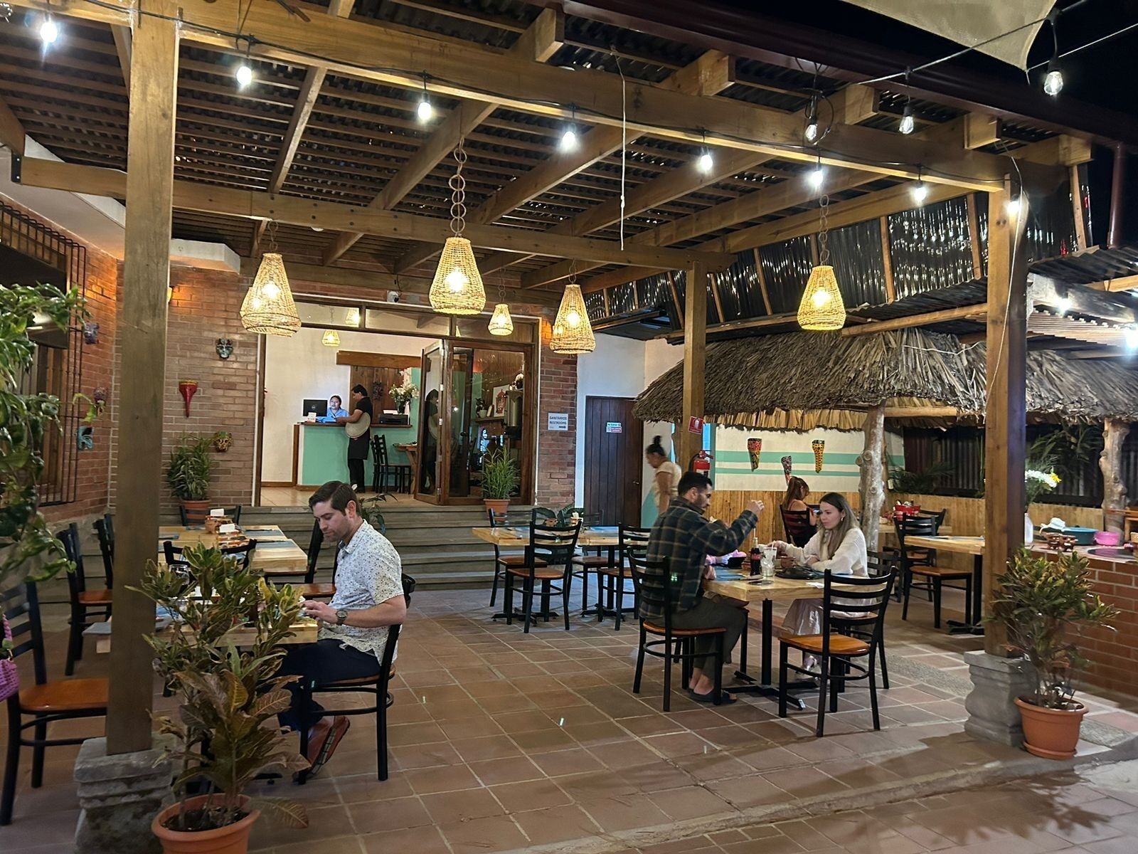 An indoor dining area with wooden beams, hanging lights, and potted plants.
