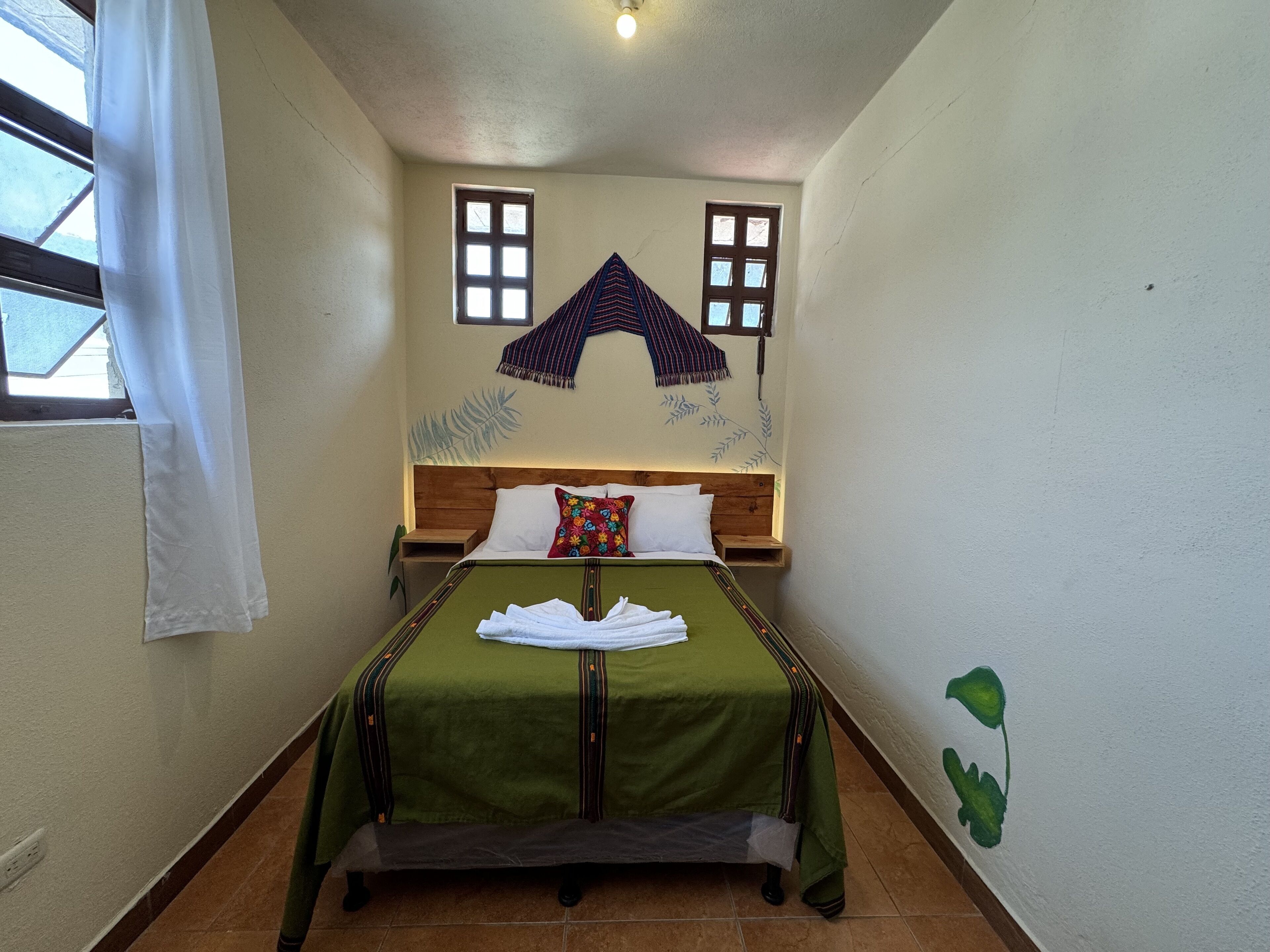 Una habitación pequeña y estrecha con una cama centrada, flanqueada por dos ventanas con marcos de madera y cortinas blancas.