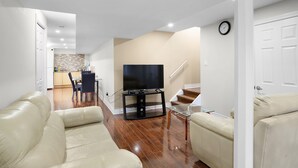Een moderne woonkamer met een flatscreen televisie op een standaard, leren banken, een eethoek met tafel en stoelen, en een trap die leidt naar een ander niveau.