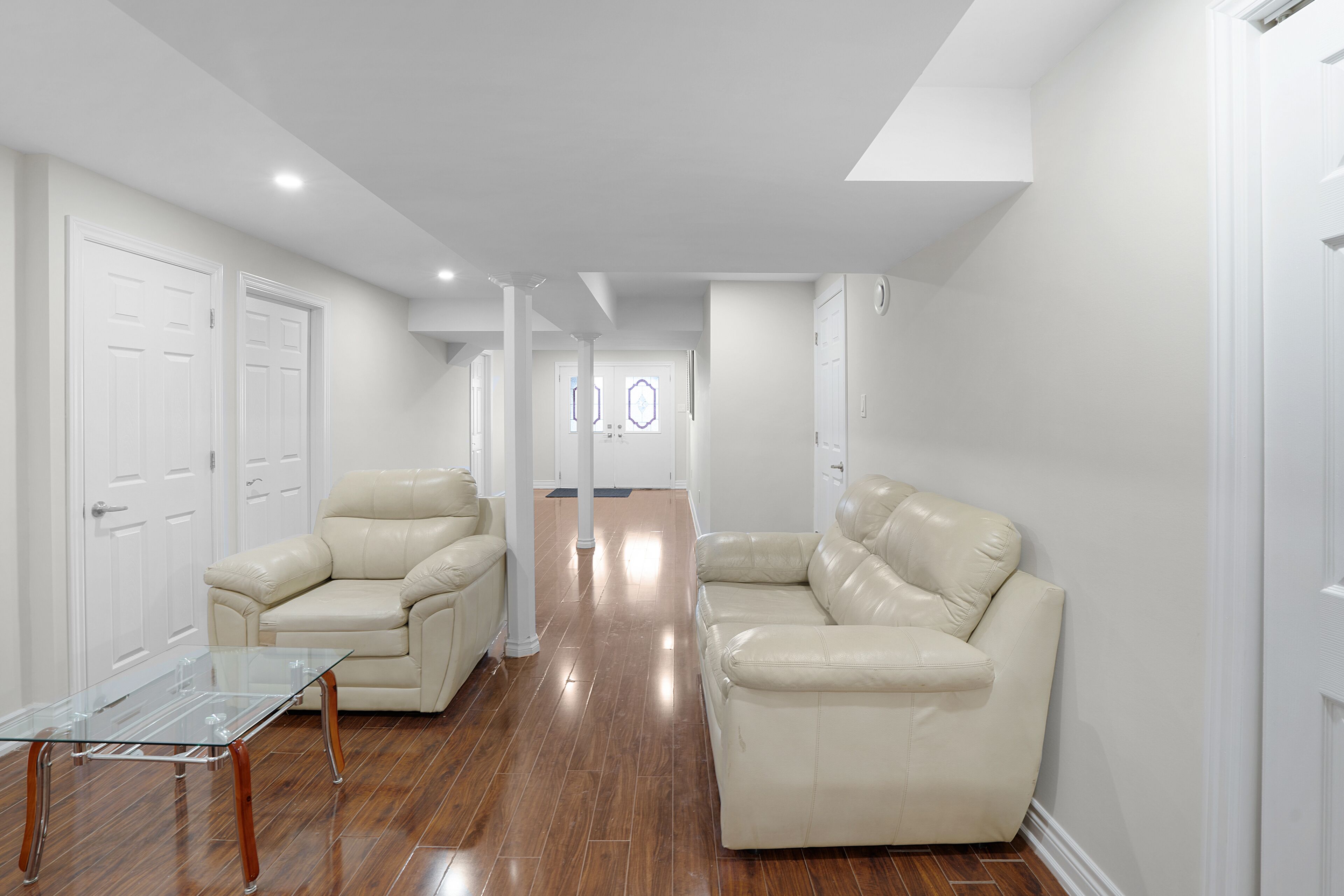 A spacious hallway with two beige leather sofas and a glass coffee table, white doors, and a wooden floor.
