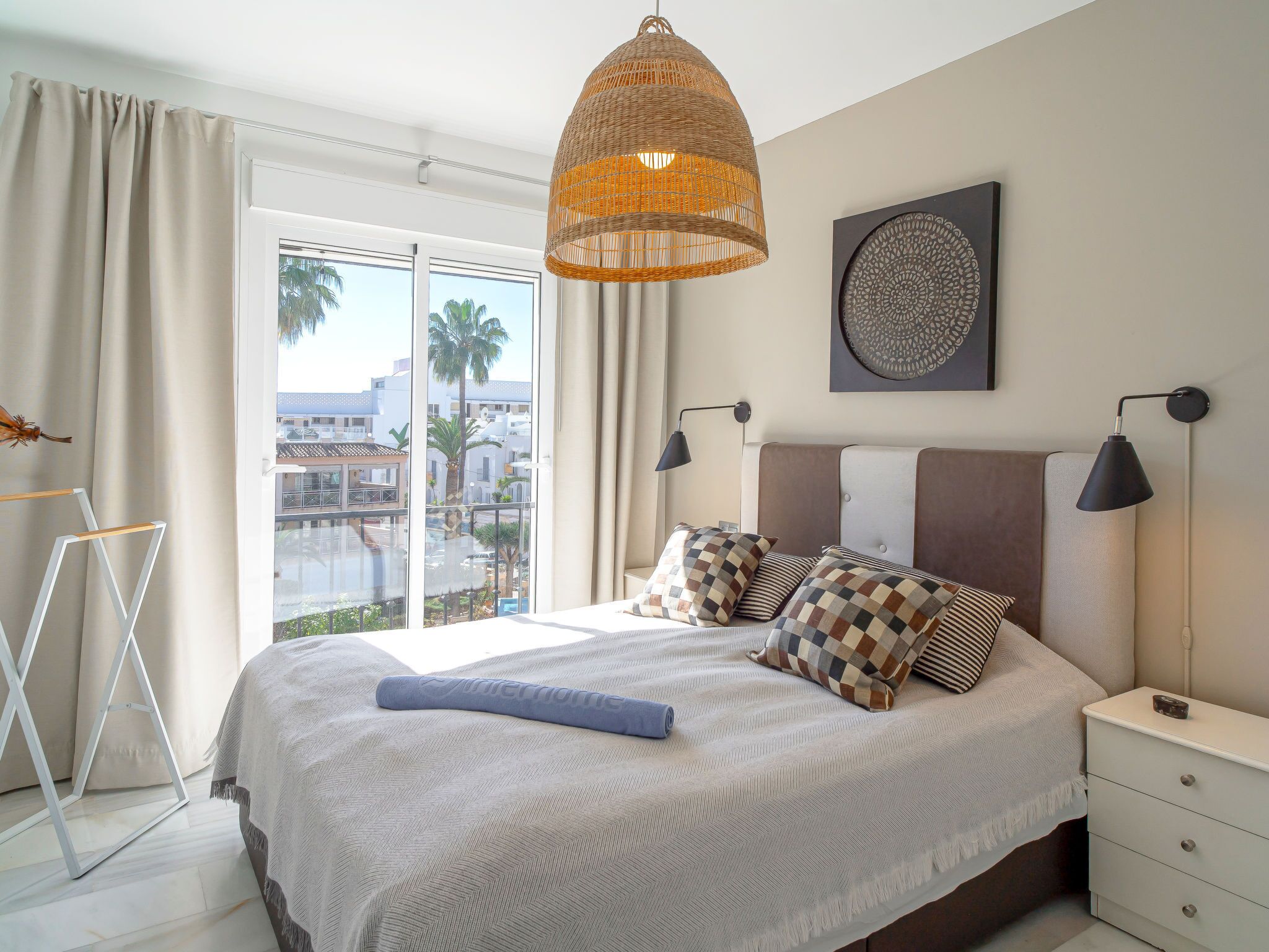 A bedroom with a large bed, a hanging wicker lamp, a nightstand, and a view of palm trees and buildings.
