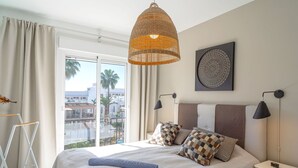 A bedroom with a large bed, a hanging wicker lamp, a nightstand, and a view of palm trees and buildings.