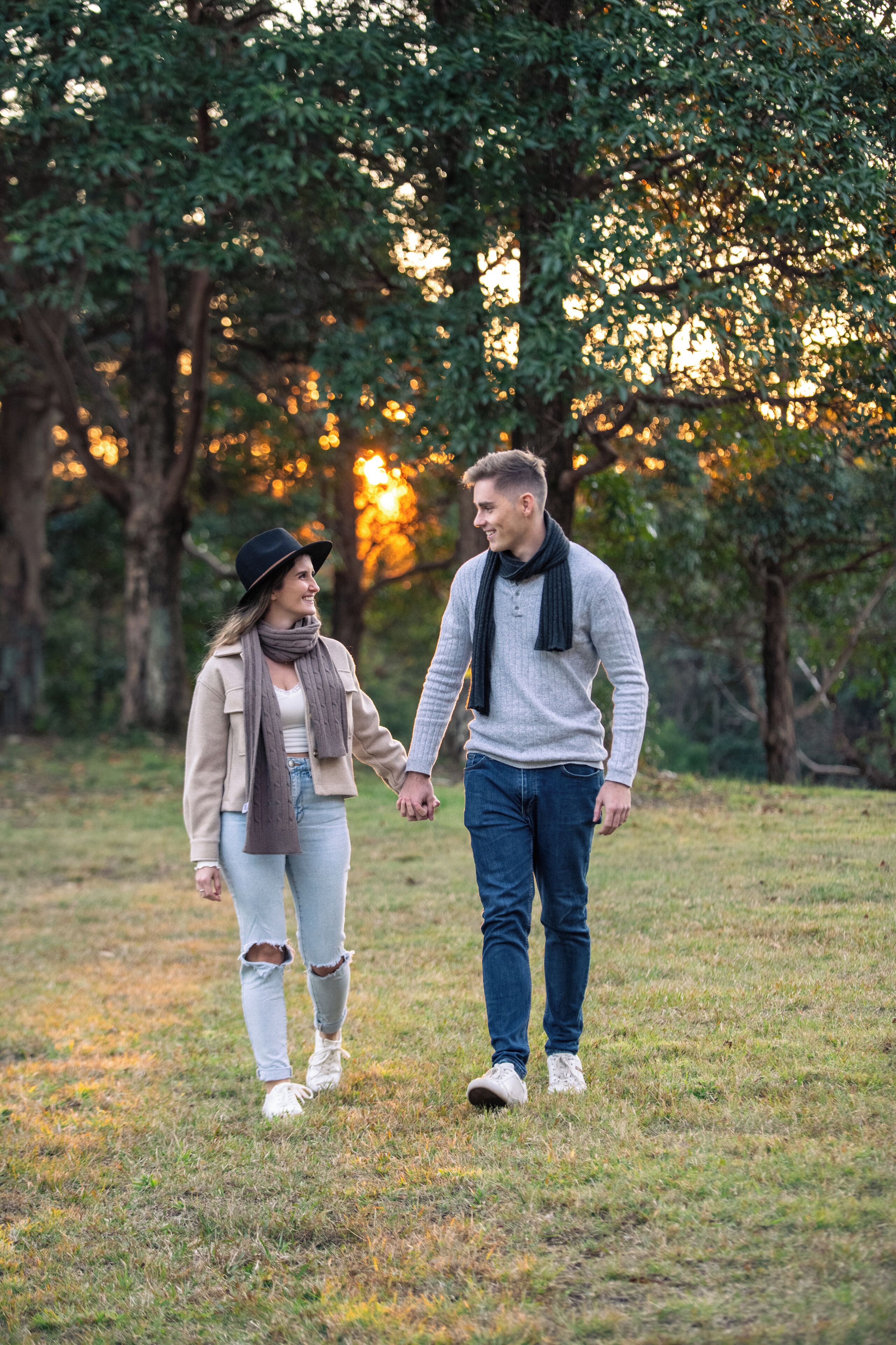 A couple walking hand in hand in a grassy park area with trees and a sunset in the background.