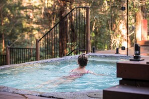 Una persona en un jacuzzi rodeado por una terraza de madera y una valla metálica, con árboles al fondo.