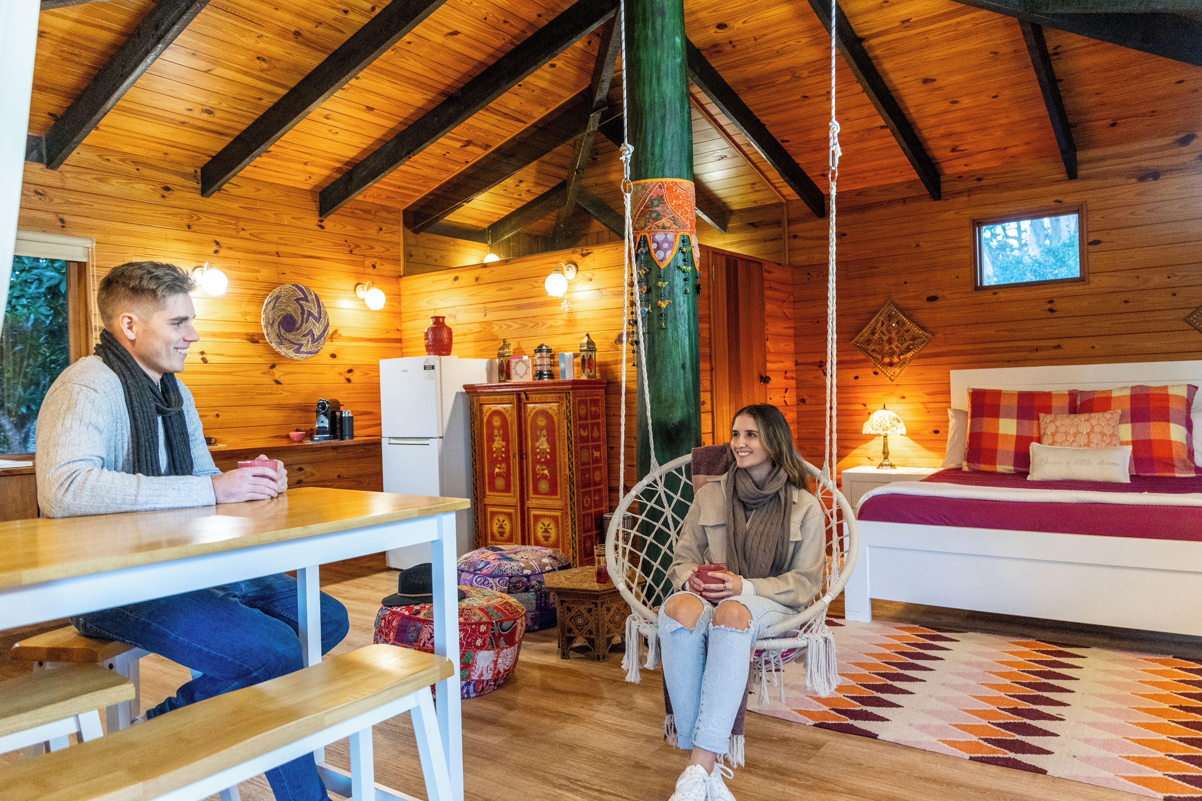 A wooden cabin interior with a dining area, a hanging chair, a bed with a red and white bedspread, and a wooden ceiling with exposed beams.