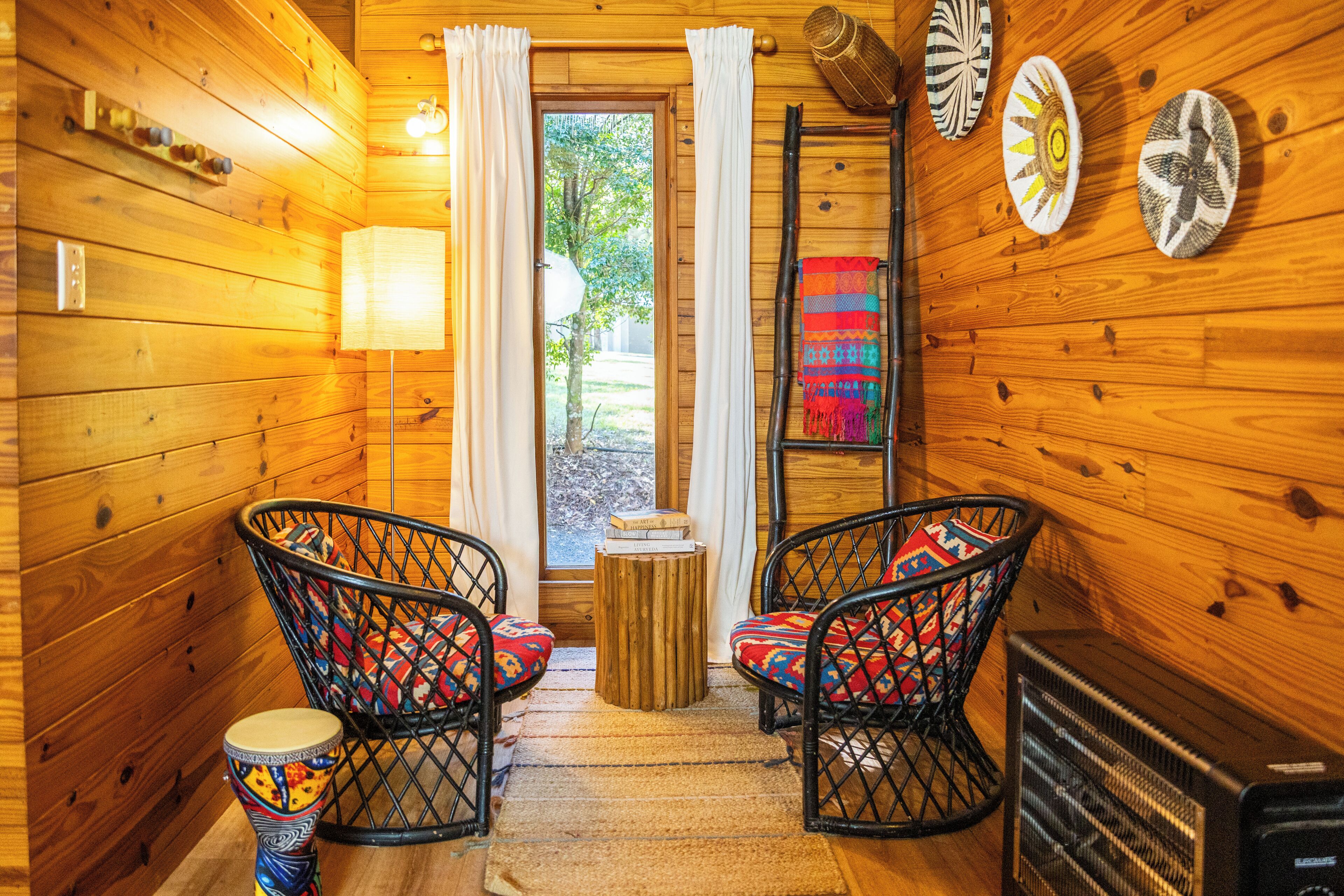 A cozy cabin interior with wooden walls, two wicker chairs with colorful cushions, a small table, and a fireplace.