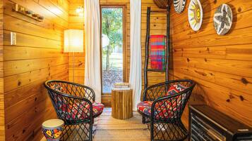 A cozy cabin interior with wooden walls, two wicker chairs with colorful cushions, a small table, and a fireplace.