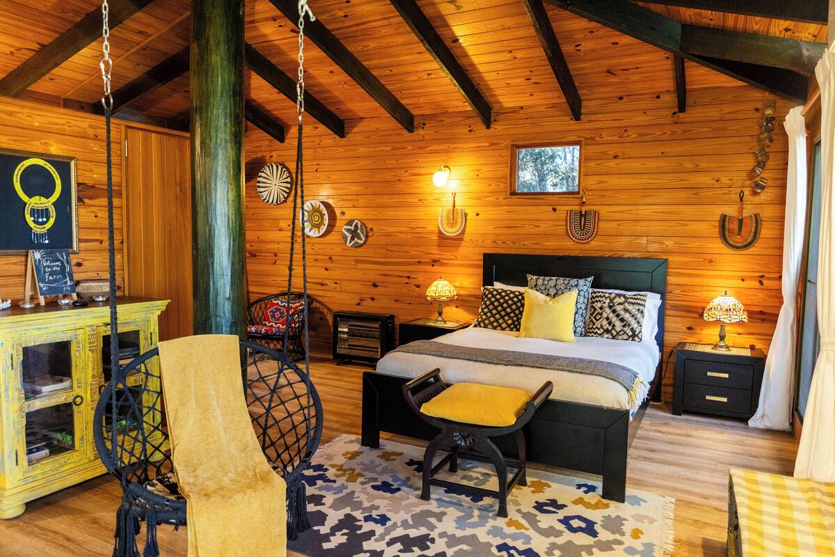 Un dormitorio rústico de cabaña de madera con cama, mesitas de noche, una silla colgante y un pequeño armario.