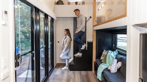 A modern interior with a staircase, a sofa with blankets, and a view through large glass doors. -  Kanimbia Cottages (Coolabine)