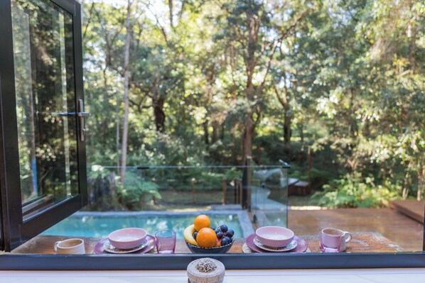 A dining area with a table set for two, a bowl of fruit, and a view of a swimming pool and forest.