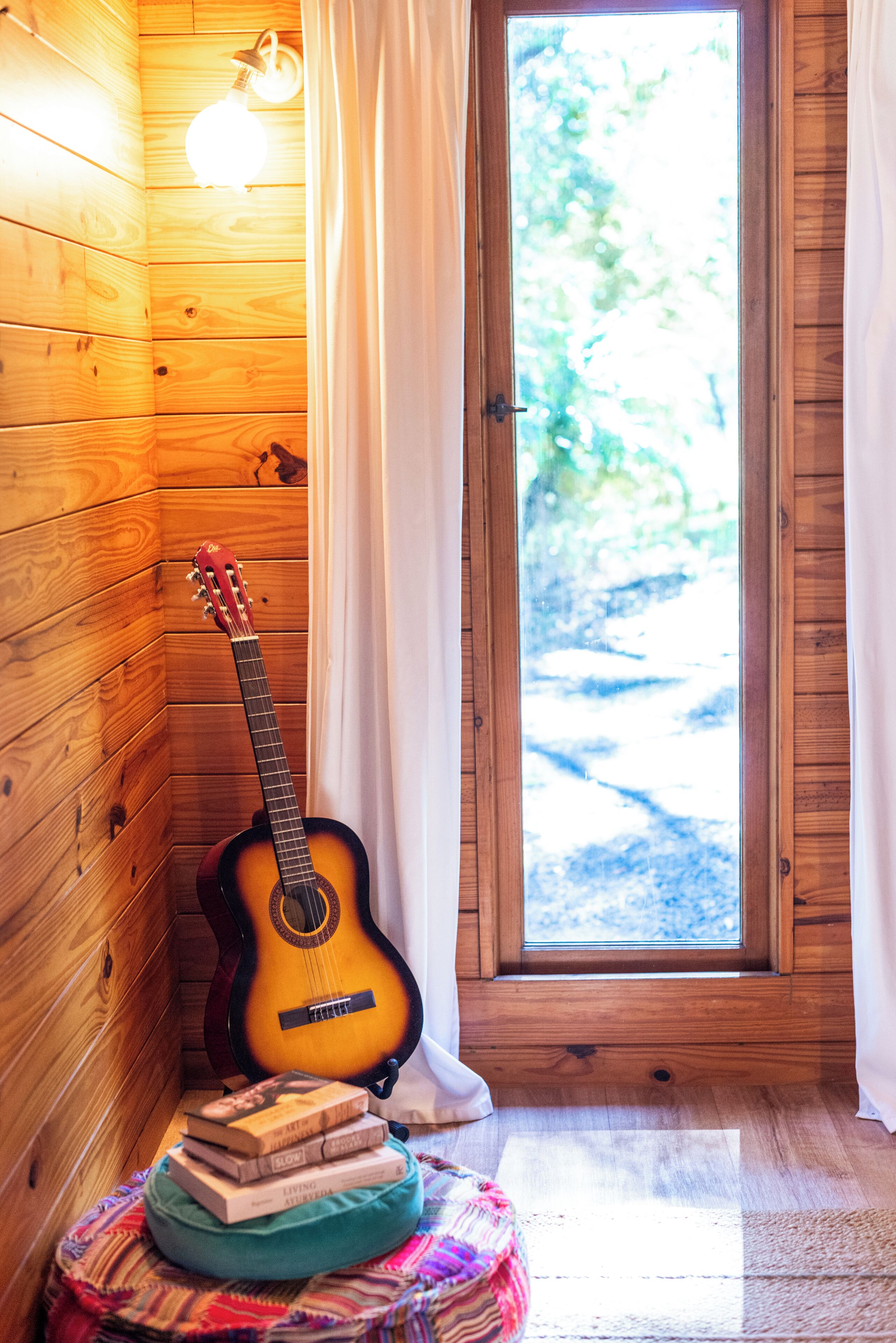 Ein Zimmer mit Holzwänden, einer Gitarre, die an der Wand lehnt, einem Fenster mit Vorhängen und einem Stapel Bücher auf einem Kissen.
