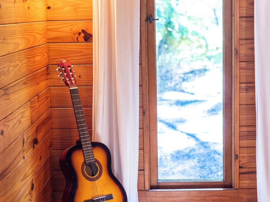 Ein Zimmer mit Holzwänden, einer Gitarre, die an der Wand lehnt, einem Fenster mit Vorhängen und einem Stapel Bücher auf einem Kissen.