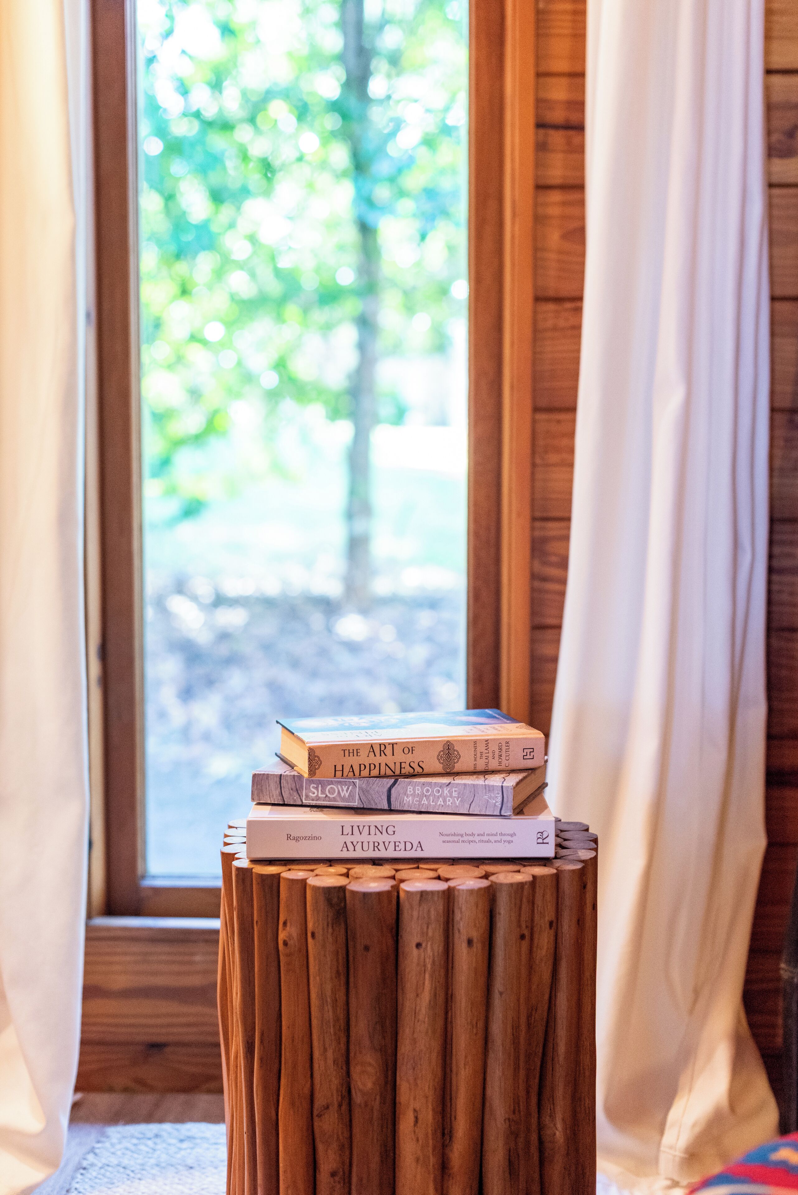 Une pile de livres posée sur un tabouret en bois près d’une fenêtre ornée de rideaux.