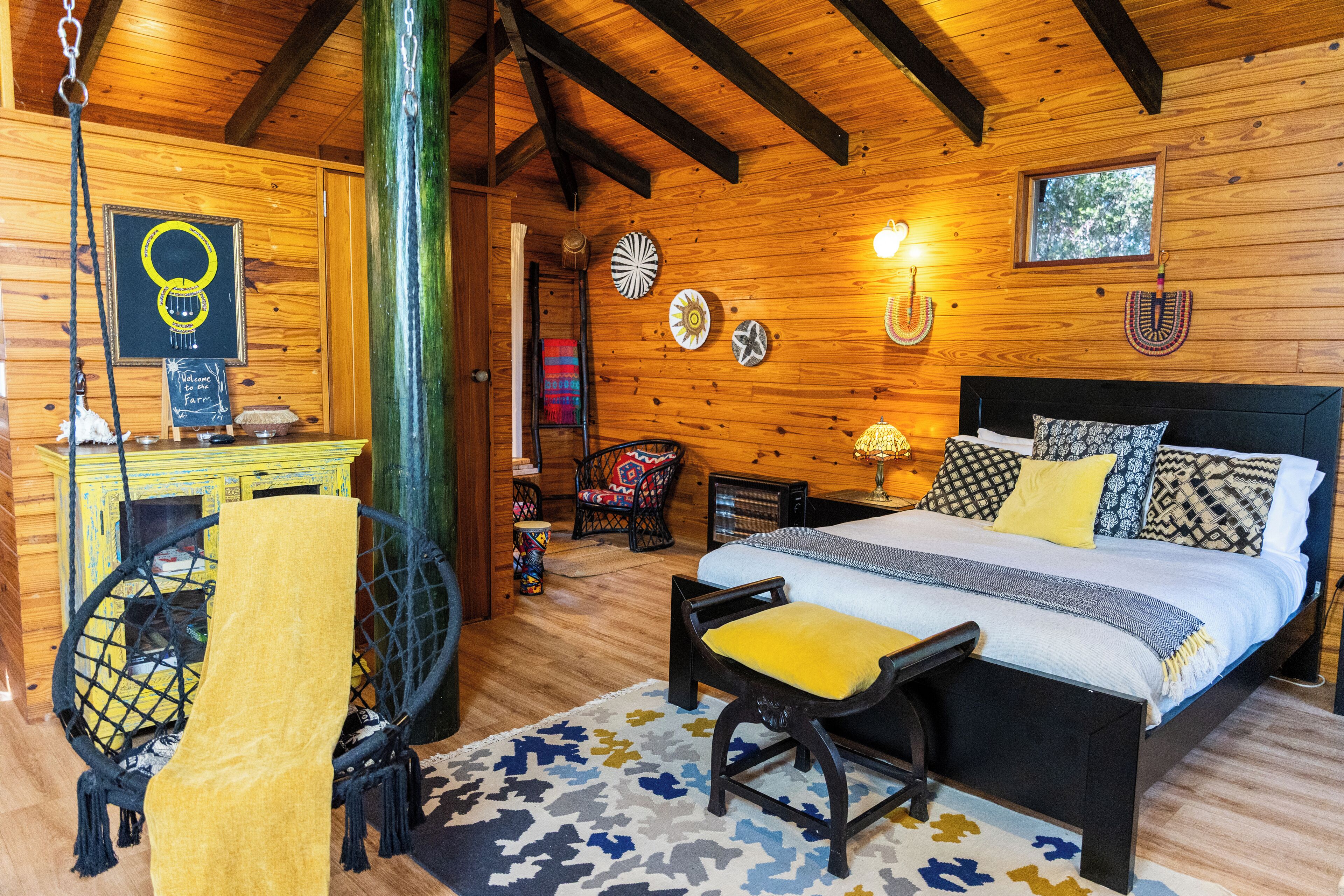 A wooden cabin interior with a bed, a chair, a hanging chair, and a small table with a chalkboard sign.
