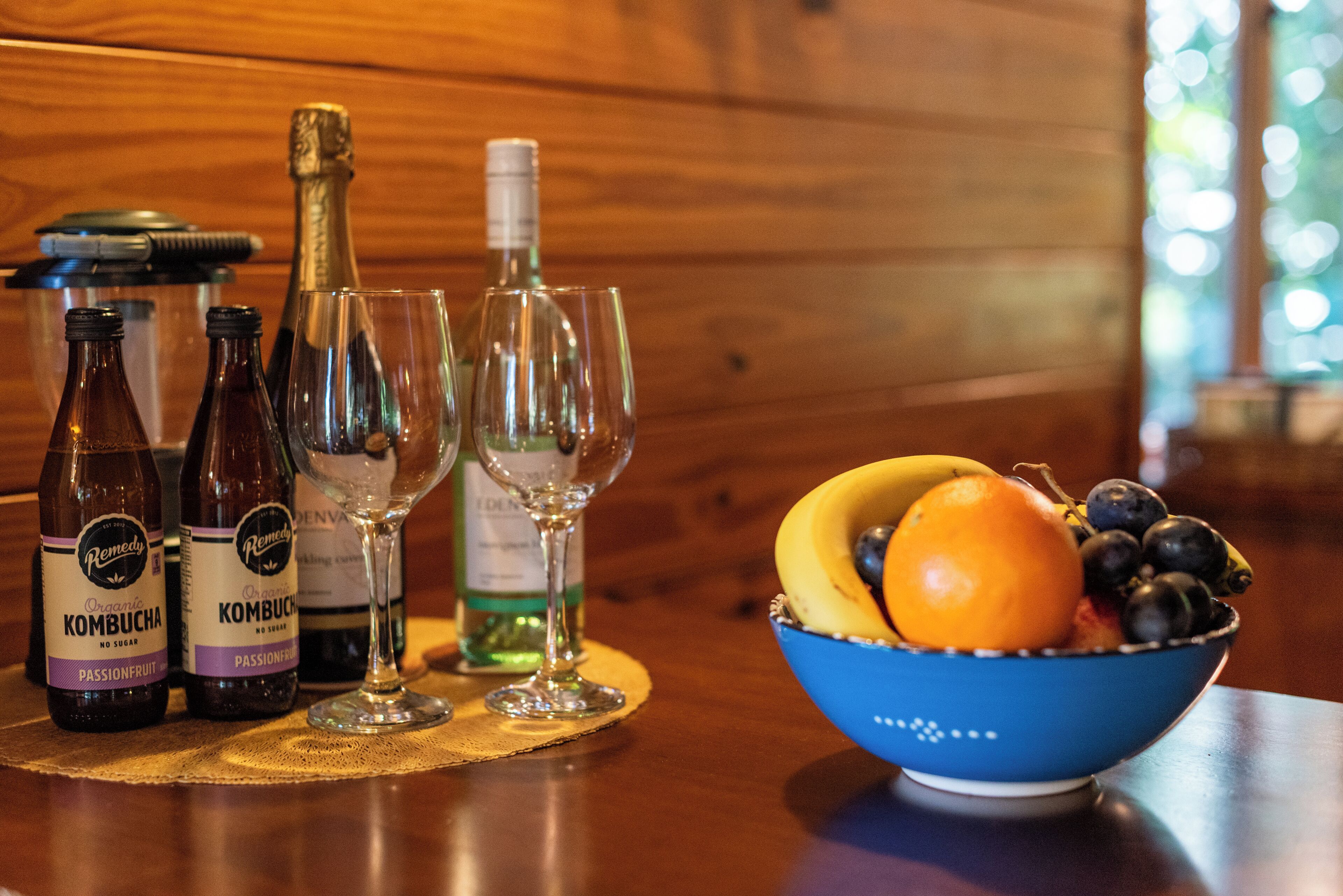 Uma mesa com garrafas de kombucha, uma taça de vinho e uma tigela de frutas.
