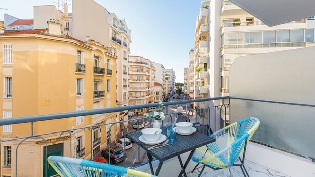 Un balcón con una mesa lista para dos personas, con vista a una calle con edificios.