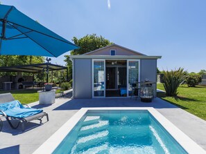 Ein Schwimmbecken mit einer Terrassenanlage, einem überdachten Sitzbereich und einem kleinen Gebäude mit einer Glasdoor.