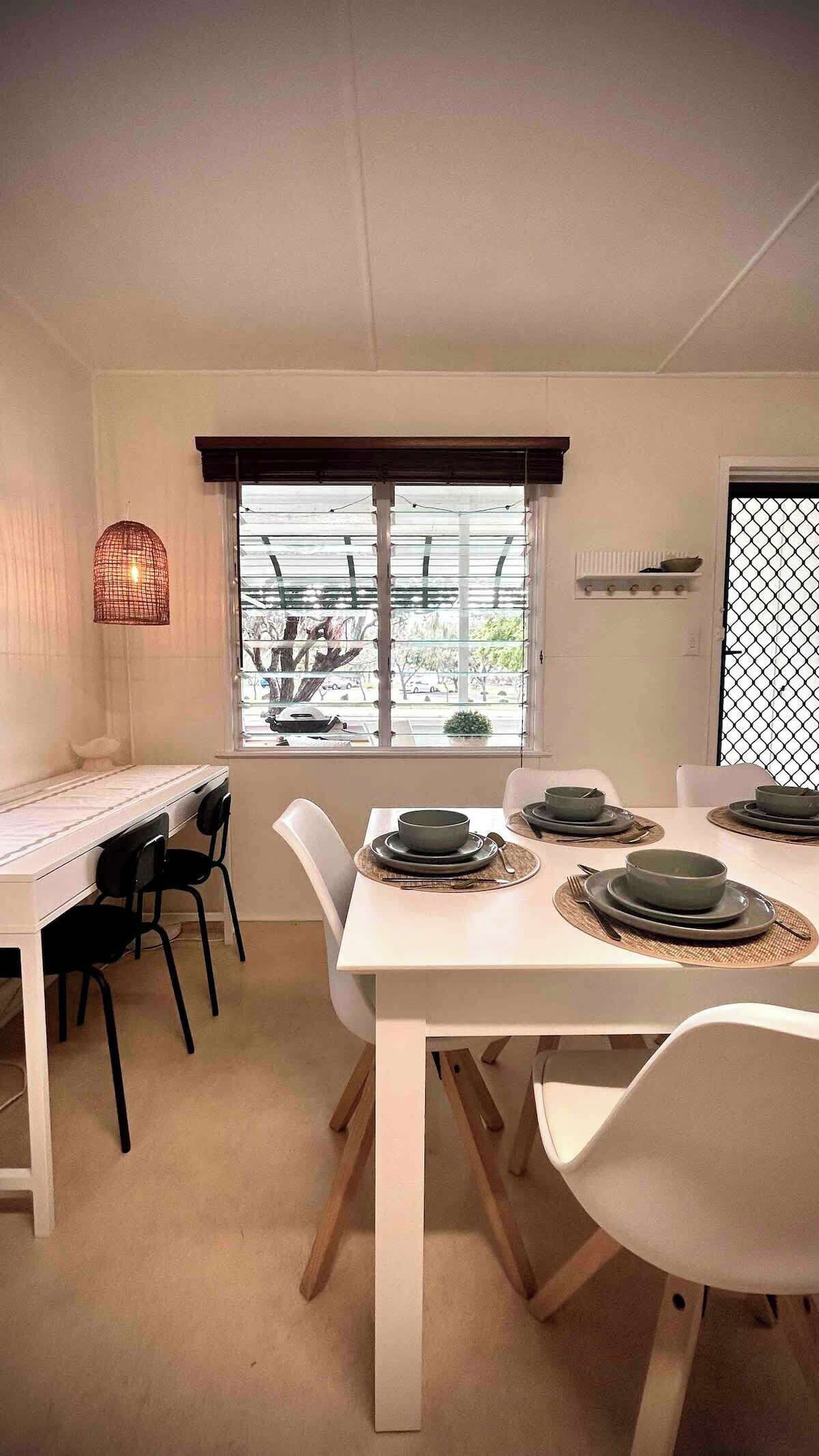 Un coin repas avec une table dressée pour quatre personnes, des chaises, une fenêtre avec des stores, un luminaire suspendu et une vue sur l’extérieur.