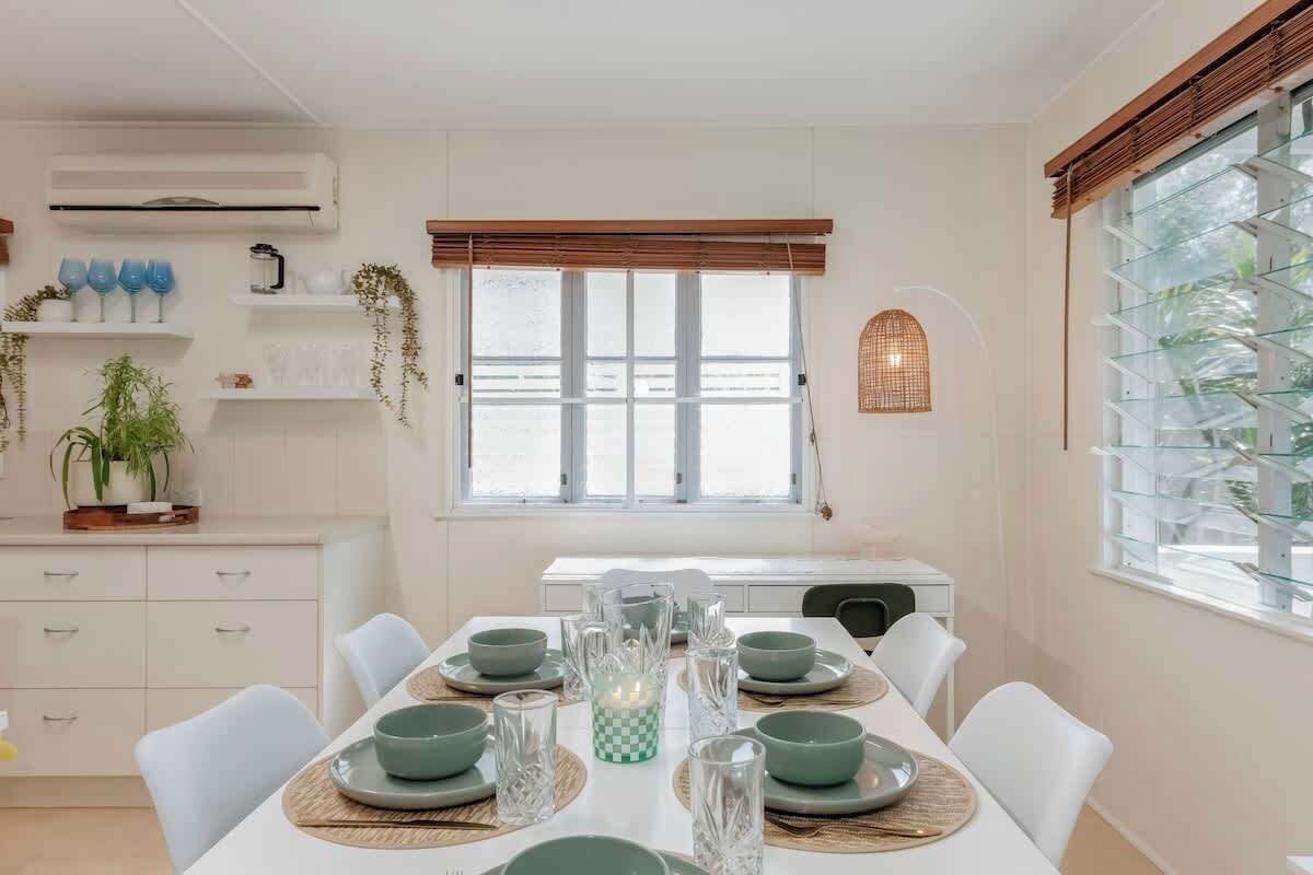 Uma sala de jantar com uma mesa posta para quatro pessoas, cadeiras brancas e uma janela com persianas.
