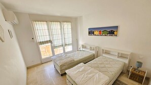Ein Schlafzimmer mit zwei Betten, einem fensterbelaubten Fenster, einer Wand-Klimaanlage und einem Bild von bunten Häusern.