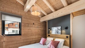 A bedroom with a wooden ceiling, a bed with white and pink bedding, a window with a view of buildings, and a wall-mounted light fixture. - L'ATTIQUE - brand new apartment for 10 people in the center of Chatel (CHATEL)