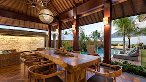 Una mesa de comedor de madera con sillas, un muro decorativo y vistas a una piscina y palmeras.