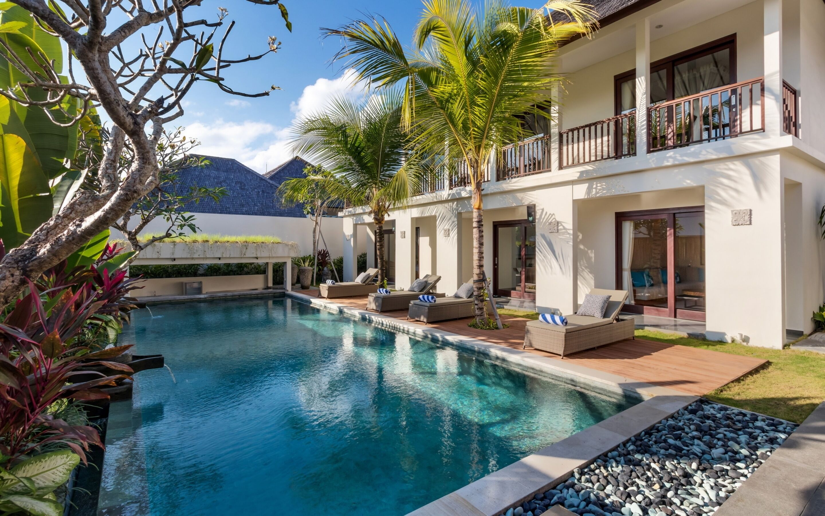 Un área de piscina moderna con sillones de descanso y una piscina cristalina, rodeada de palmeras y un edificio blanco con balcones.