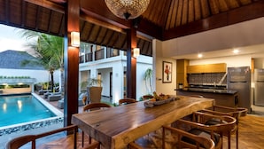 Una cocina moderna con estufa y refrigerador integrados, un comedor con mobiliario de madera, y un espacio de descanso junto a la piscina con vistas a ella.