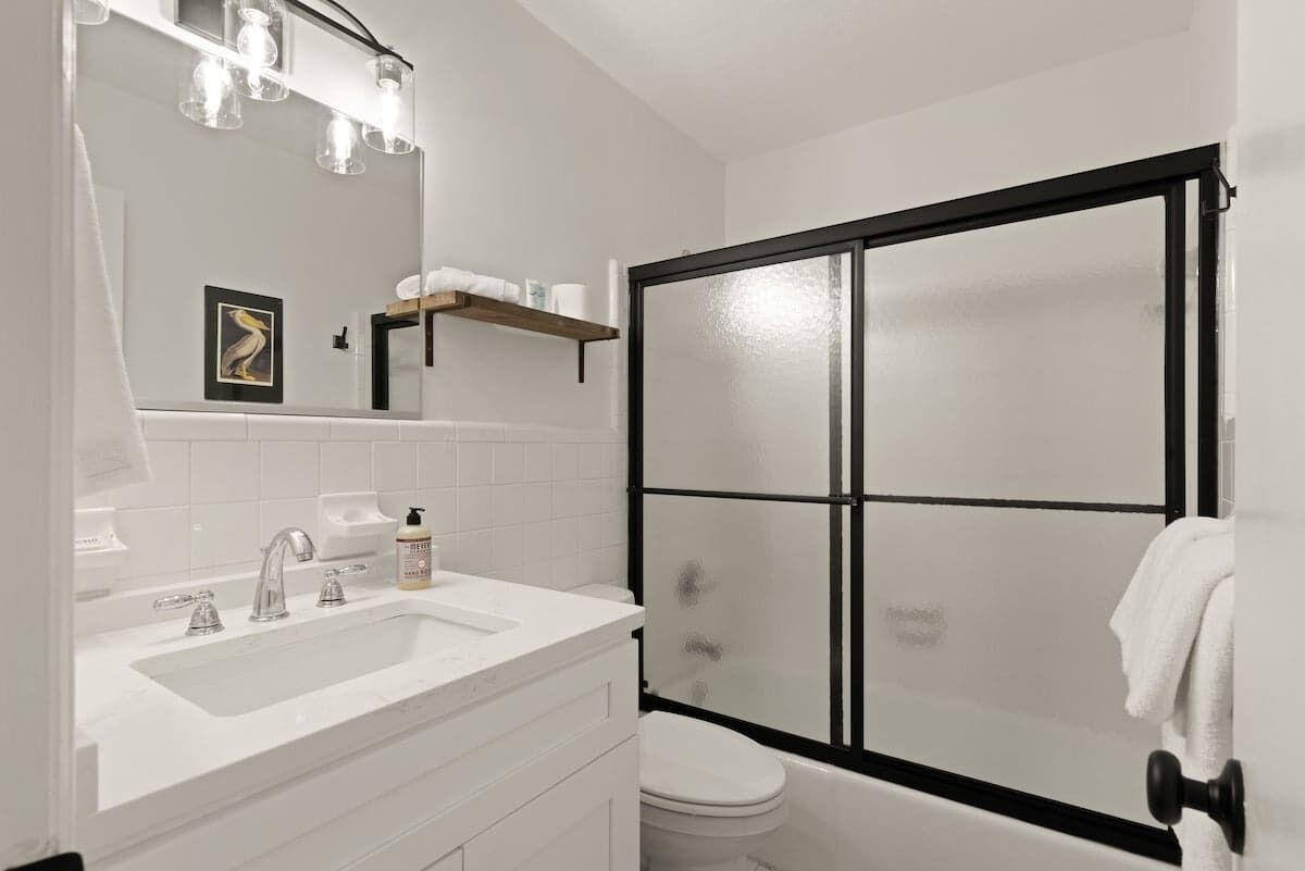 Un baño con lavamanos blanco, espejo e inodoro. Hay una ducha con marco negro y puertas de cristal satinado.
