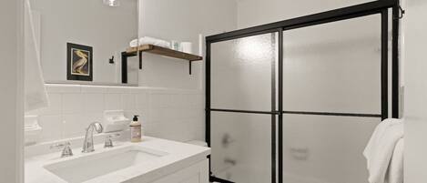 Un baño con lavamanos blanco, espejo e inodoro. Hay una ducha con marco negro y puertas de cristal satinado.