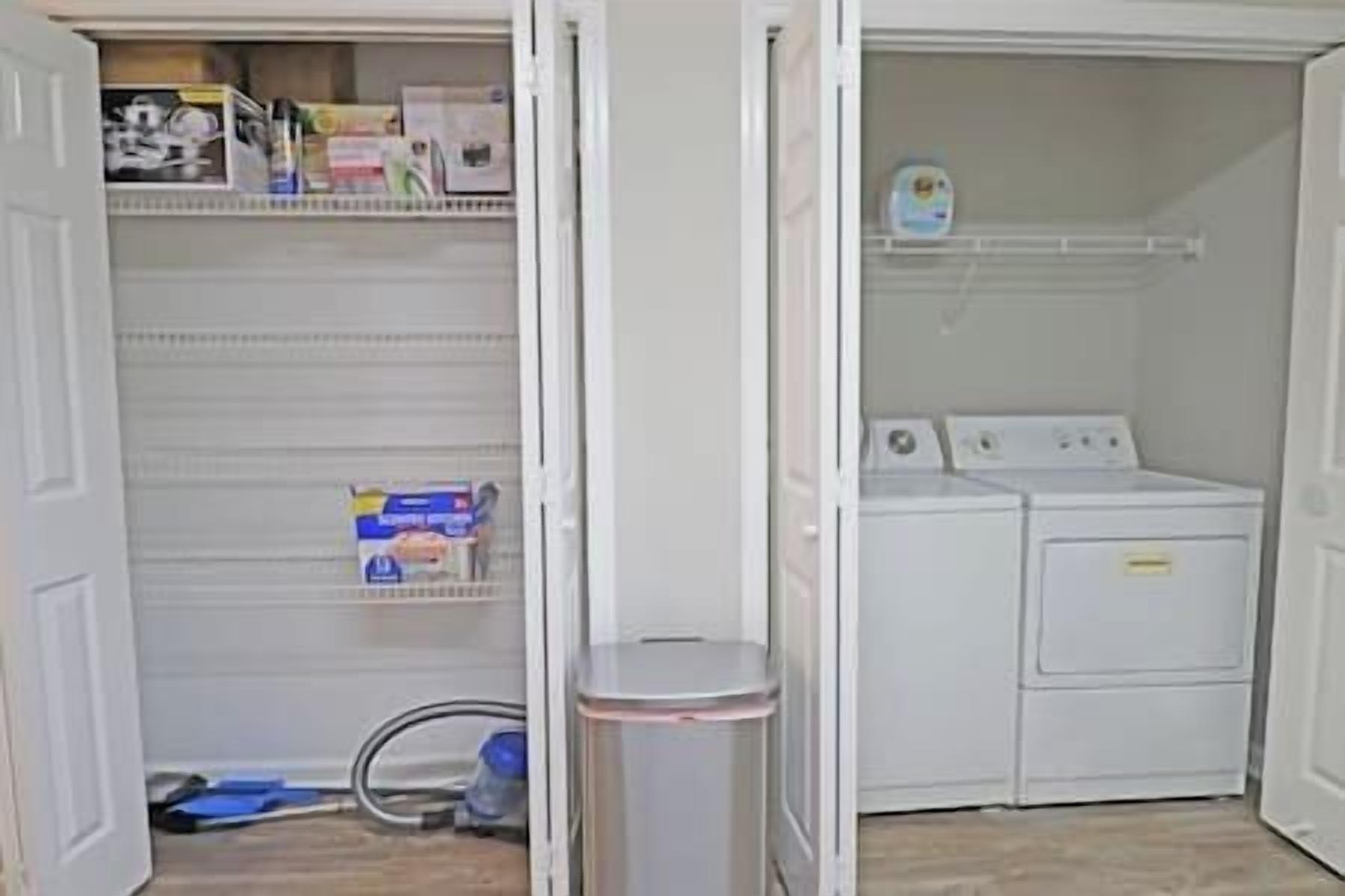 A laundry room with a washing machine and dryer, a trash can, and shelves with cleaning supplies.