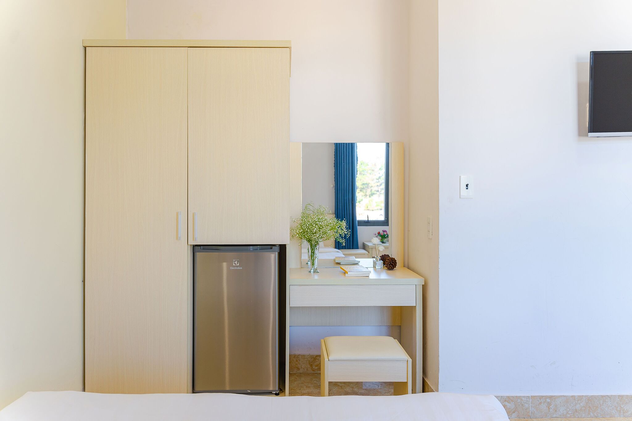 Habitación de hotel pequeña con cama, un escritorio con espejo, un refrigerador y ventana con cortinas azules.
