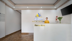 A reception desk with a logo, a woman standing behind it, and a plant on the counter. - RAON Central Hotel (Xuan Huong - Da Lat)
