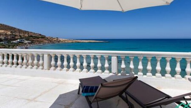 En balkon med hvid gelænder, en hvid parasol og to liggestole med udsigt over kysten.