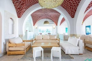 A spacious room with a large bed, two white armchairs, and a small table. The room features a high, arched ceiling with a woven design and brick accents. - Dar Barsha Beach Front White Sand and Happiness (Raf Raf)